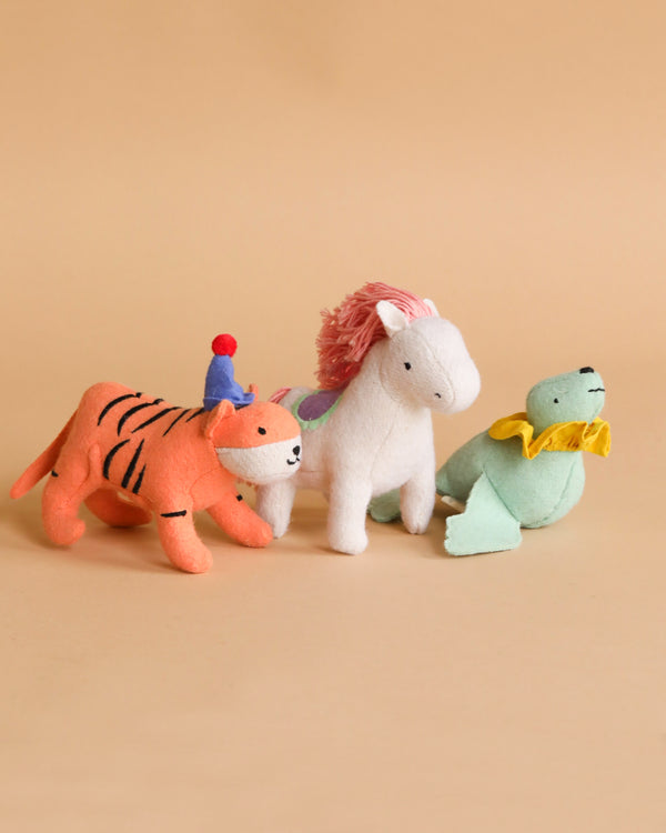 A group of three Olli Ella Felt Holdie Circus Animals are arranged on a beige background. From left to right: an orange tiger wearing a blue party hat, a white horse with a pink mane and a colorful saddle, and a green frog with a yellow ruffled collar. These handmade wool toys inspire imaginative play.