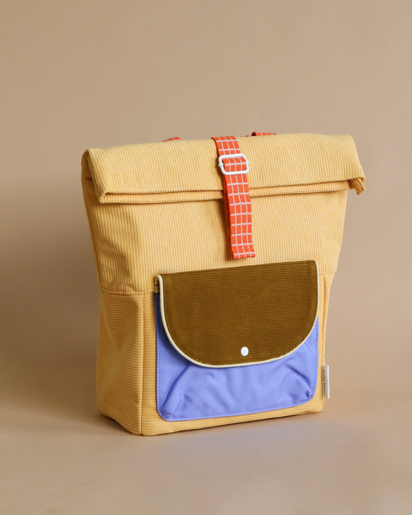 Sticky Lemon corduroy backpack with a front blue pocket, secured by a beige strap with a red and white square pattern. The background is a neutral beige.