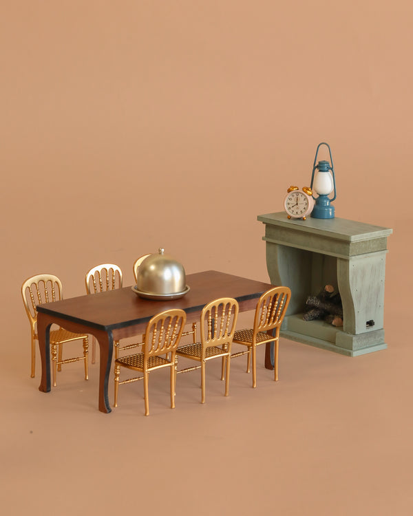 The Maileg Grand Dining Room Set includes a dining table, six gold chairs, a covered serving dish, and a mantelpiece with clock and lantern—perfect for Maileg Mouse Size figures in any dollhouse.