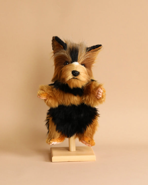 A small, hand-sewn Yorkie Terrier Dog Puppet with fluffy brown and black fur is positioned upright on a wooden stand against a beige background. The toy, resembling HANSA animals, has a cute, realistic appearance with pointed ears and a soft, textured body.