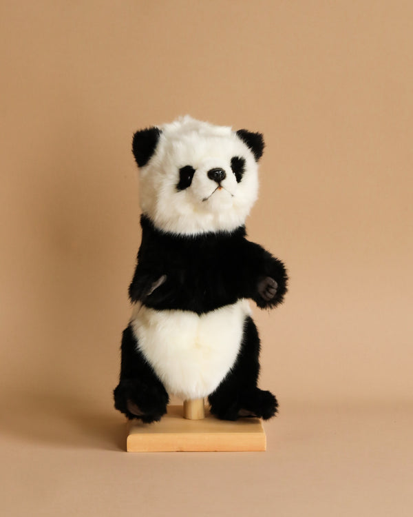 A small Panda Puppet stands on a light wooden base against a plain beige background. The toy, part of a range of realistic plush toys, features a classic panda color pattern with black arms, legs, ears, and eye patches, complemented by white fur on the rest of its body.