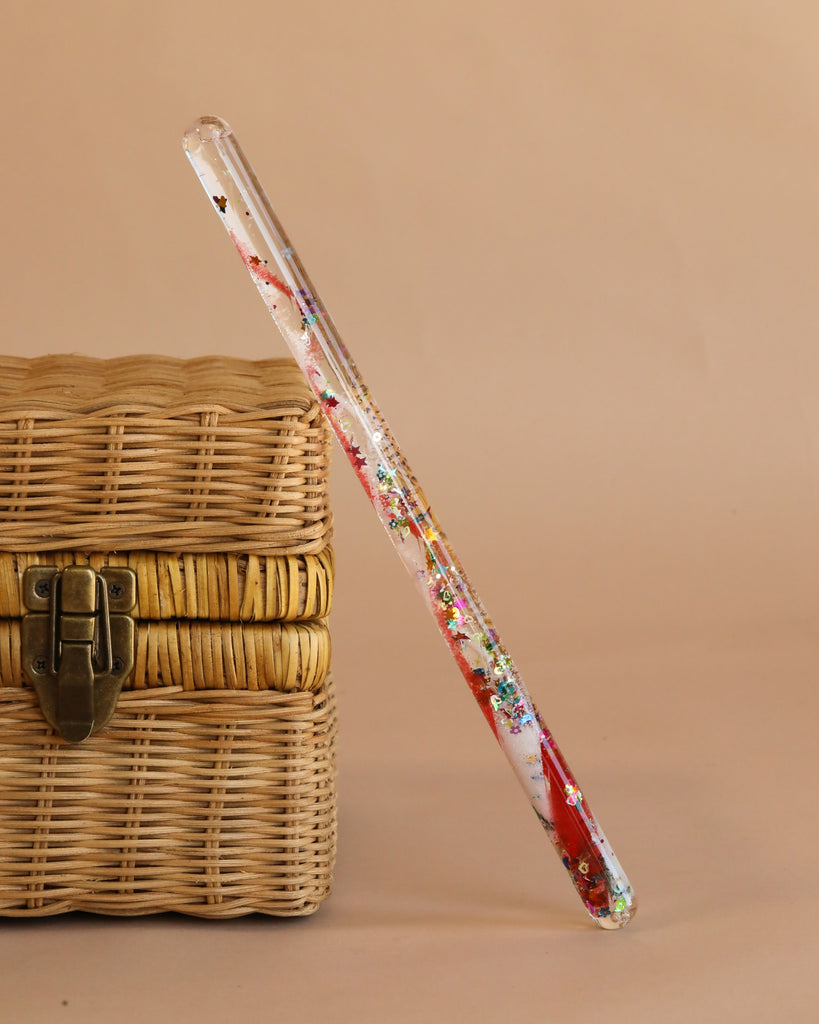 The Spiral Glitter Magic Wand - Red, a clear resin dip pen with red, white, gold accents and rainbow glitter details, stands upright against a closed woven basket with a brass latch on a plain beige background.