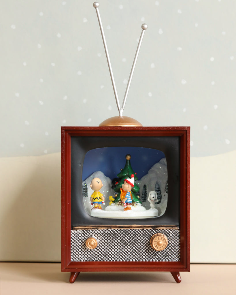 The Charlie Brown Christmas Music TV features a vintage-style design that illuminates a scene of two cartoon characters outside in winter beside a Christmas tree. Complete with antennas and knobs, this TV plays a soft Christmas melody while the pale background is speckled to resemble falling snow.