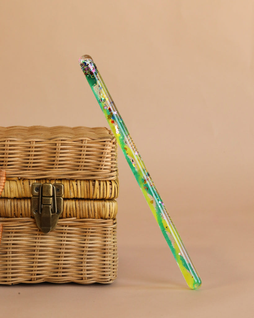 A Spiral Glitter Magic Wand - Green/Yellow filled with confetti and rainbow glitter stands upright next to a closed wicker box with a metal latch against a beige background—a perfect imagination toy for kids.