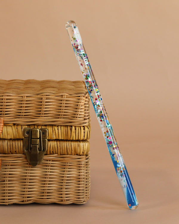 The Spiral Glitter Magic Wand - Blue, with blue tips and colorful glitter details, rests against a woven wicker basket with a metal latch, set on a plain beige background—a magical toy inspiring kids' imagination.