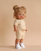 The Minikane Standing Doll (14") - Louisa with Clothing features light skin, curly blonde pigtails, and blue eyes. She stands against a beige background wearing an anatomically correct beige knit outfit and white sneakers with animal print, made in Spain.