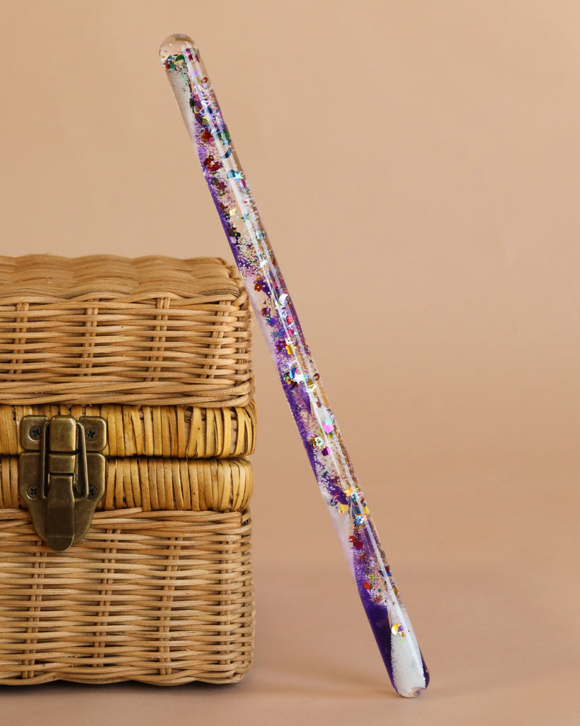 A Spiral Glitter Magic Wand - Purple, filled with colorful glitter and decorations, leans against a closed wicker basket with a brass latch on a beige background—an enchanting toy for imaginative kids.