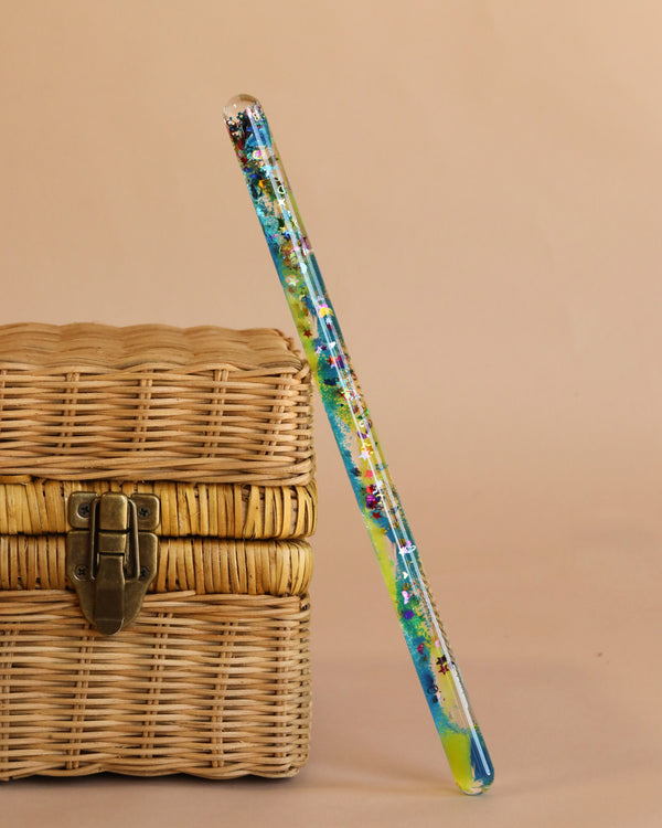 The Spiral Glitter Magic Wand - Blue/Yellow, with its colorful glass design and confetti-like shimmer, rests against a closed wicker basket with a metal clasp on a plain beige background.