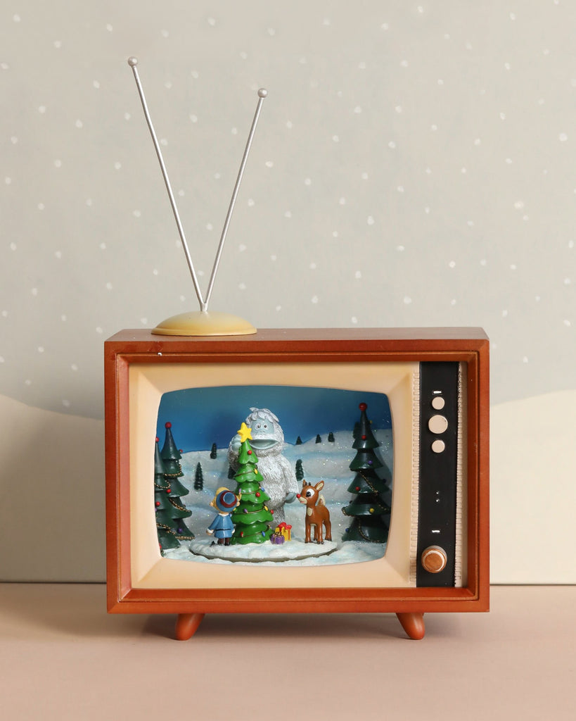 The "Christmas Music TV With Rudolf" features a retro television set that lights up to display a charming winter scene with a snow monster, Rudolph the Red-Nosed Reindeer, and Christmas trees. It comes with an antenna on top and boasts a wooden exterior, all set against a snowy backdrop on a beige surface.