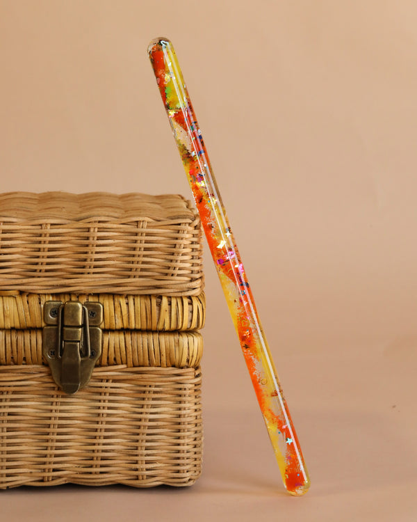 The Spiral Glitter Magic Wand - Orange, featuring rainbow glitter, floral decorations in orange, yellow, and red, and silver stars, rests against a closed wicker basket with a metal latch on a beige background.