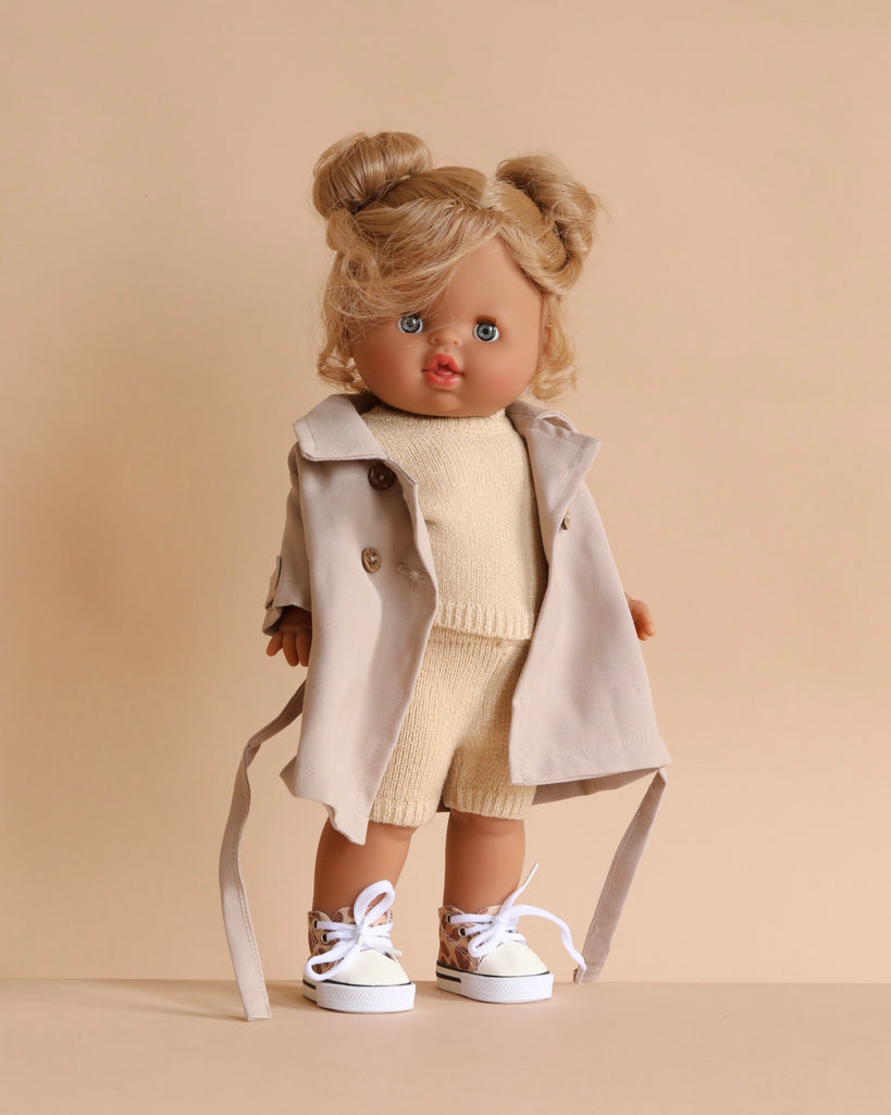 The Minikane Standing Doll (14") - Louisa with Clothing features a blonde doll with blue eyes in two buns, dressed in a light beige coat, knit shorts and top, and white sneakers against a plain beige background.