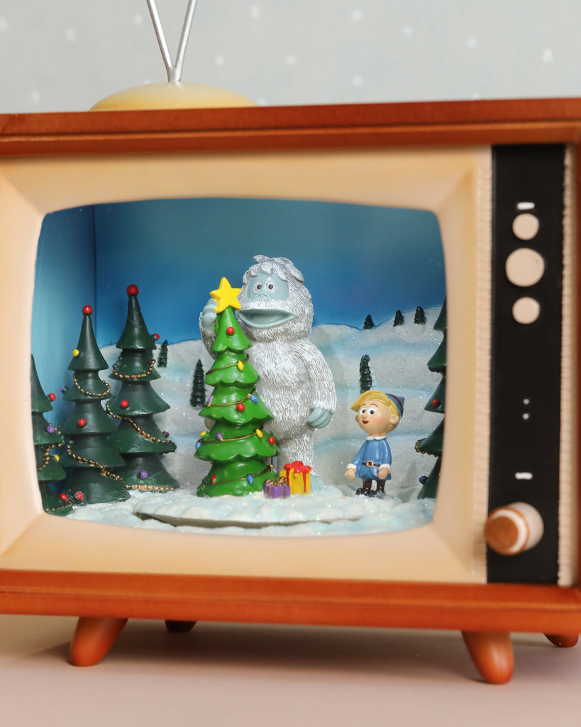 Housed in a retro TV, the Christmas Music TV With Rudolf diorama showcases a winter scene where a large white creature and small boy stand beside a festively decorated Christmas tree. The setting is surrounded by snow and pine trees, with colorful presents at the base, capturing the nostalgic charm of Rudolph The Red-Nosed Reindeer. Soft volume-controlled lights enhance the ambiance.