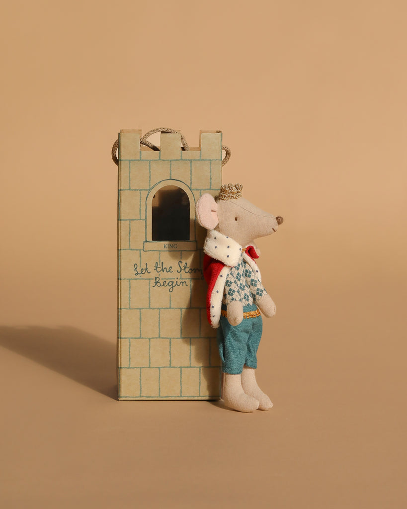 A Maileg Queen Mouse in a crown, red cape, blue pants, and floral shirt rests by the Maileg Royal Tower Set featuring a window and "Let the story begin" inscribed on it.