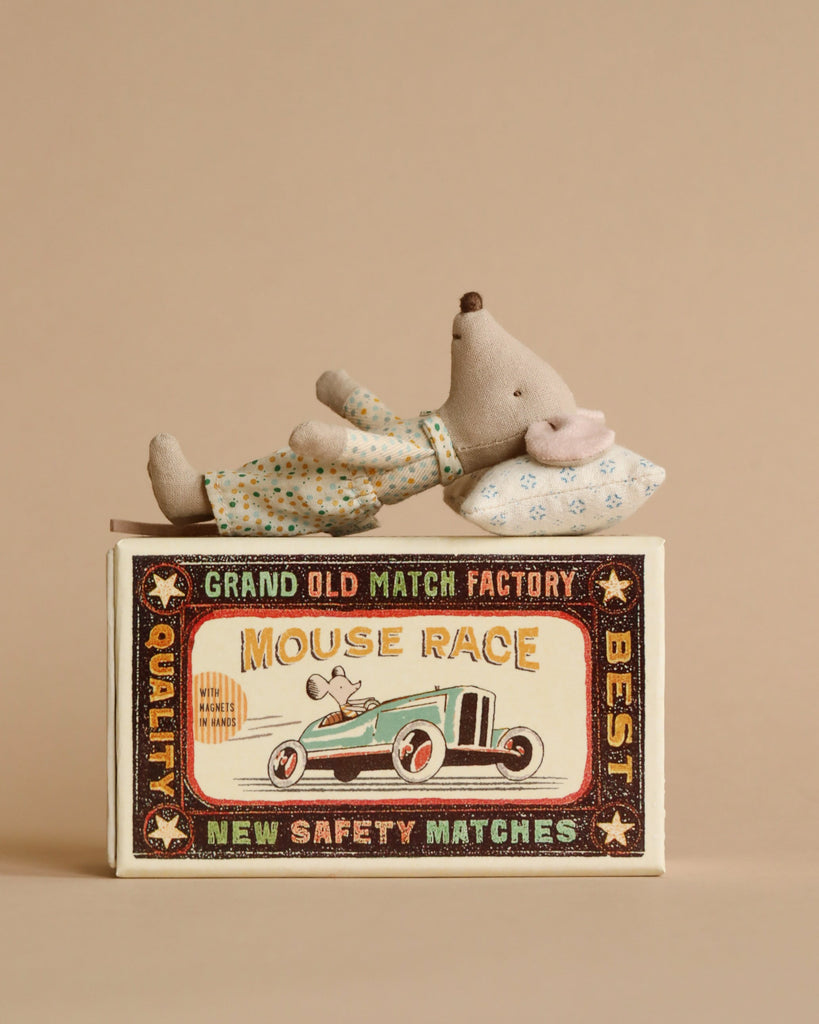 A small Maileg Little Brother mouse in matchbox toy lying on top of a colorful vintage matchbox bed with the words "Grand Old Match Factory Mouse Race." The box features an illustration of a racing car and says "New Safety Matches.
