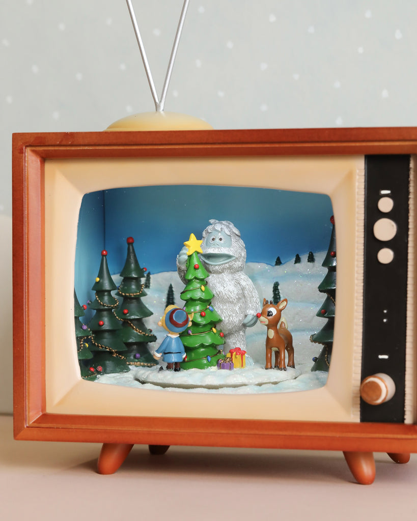 The Christmas Music TV With Rudolf showcases a vintage television set complete with volume control. It displays a winter scene featuring a snowman, a child in a blue coat and hat, Rudolph the Red-Nosed Reindeer, and Christmas trees adorned with presents. A star rests atop the central tree against a snowy backdrop and blue sky.