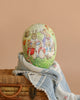 The Easter Basket Gift Set features a Maileg Easter Egg with rabbits in clothing, displayed on a woven basket draped with a blue-and-white blanket against a plain beige background.
