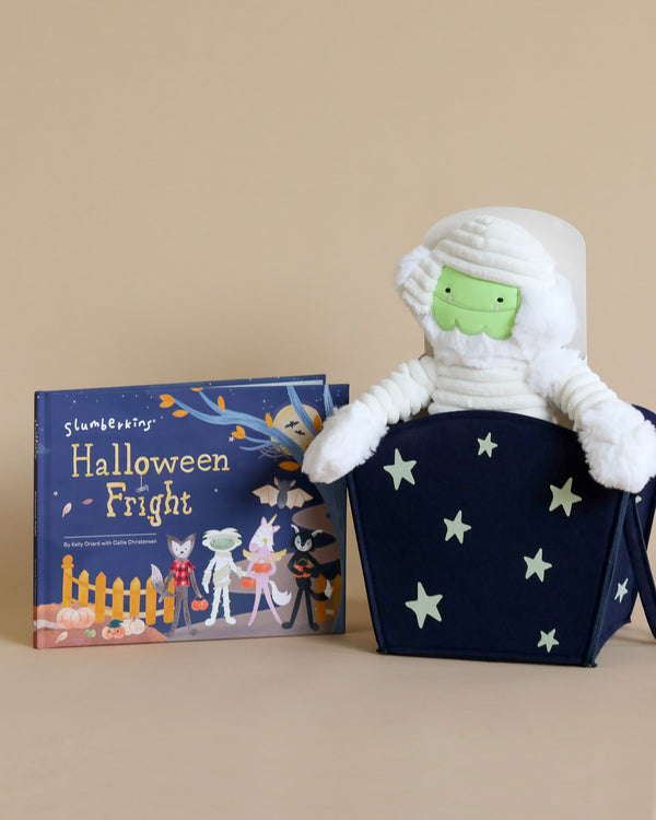 A Slumberkins Halloween Gift Set - Mummy Kin + Halloween Fright Book sits in a dark blue, star-patterned box next to a children's book titled "Halloween Fright" with illustrations