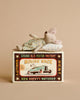 A small toy mouse in pajamas and a pillow rests on a vintage matchbox labeled “Mouse Race,” featuring an illustration of a mouse driving a car—part of the charming Maileg Family Celebration Set, perfect for expanding your Maileg accessories collection or gifting as an enchanting addition to any Maileg mouse set.