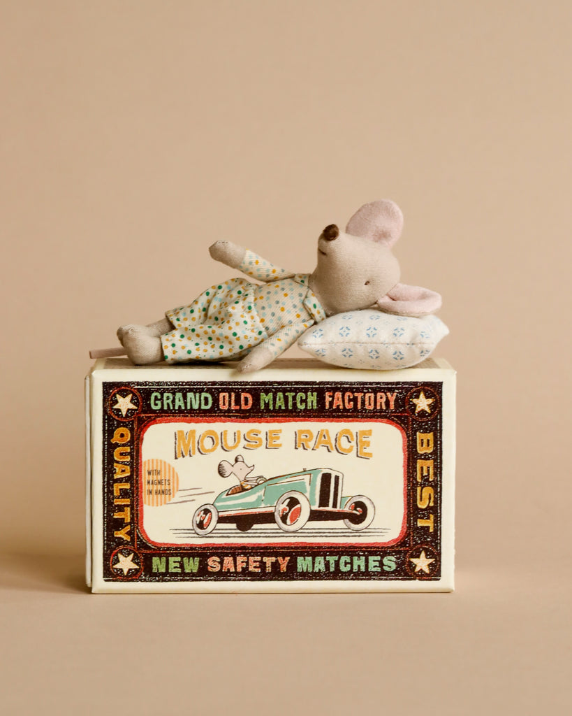 A small toy mouse in pajamas and a pillow rests on a vintage matchbox labeled “Mouse Race,” featuring an illustration of a mouse driving a car—part of the charming Maileg Family Celebration Set, perfect for expanding your Maileg accessories collection or gifting as an enchanting addition to any Maileg mouse set.