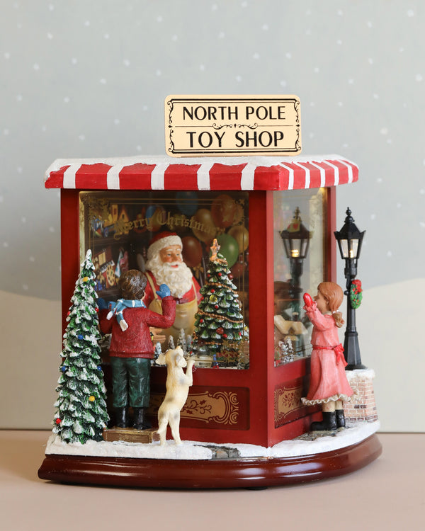 The North Pole Toy Shop With Moving Train - Musical creates a festive scene with Christmas melodies filling the air. Inside the decorative shop, which features red and white striped awnings, a Santa figure can be seen. Outside, a girl and boy stand with a reindeer amid snow-covered Christmas trees and an illuminating lantern, adding to the holiday display.