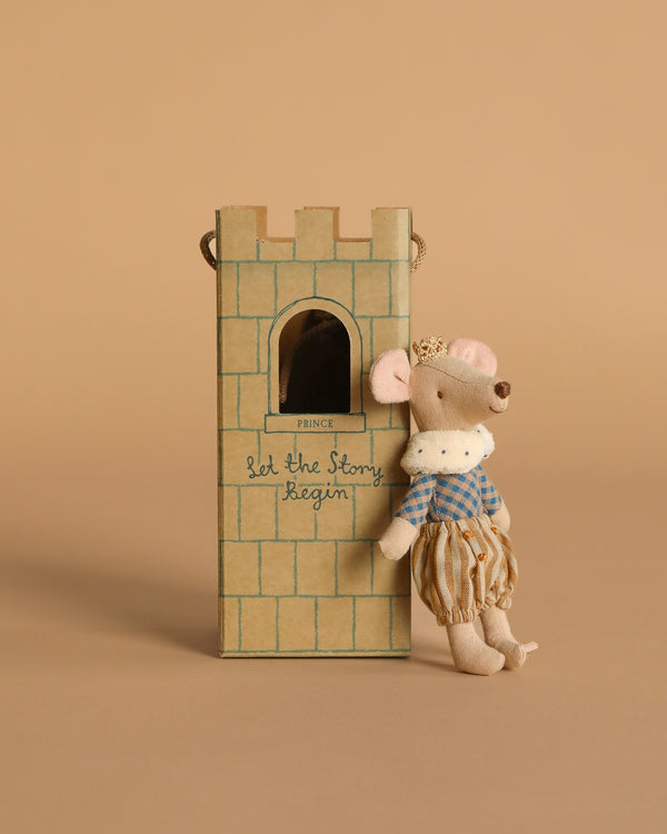 The Maileg Prince Mouse, Big Brother—a small cotton mouse in royal clothes and a crown—leans on a cardboard castle tower with a window and "Let the Story Begin" written on it. The background is light brown.