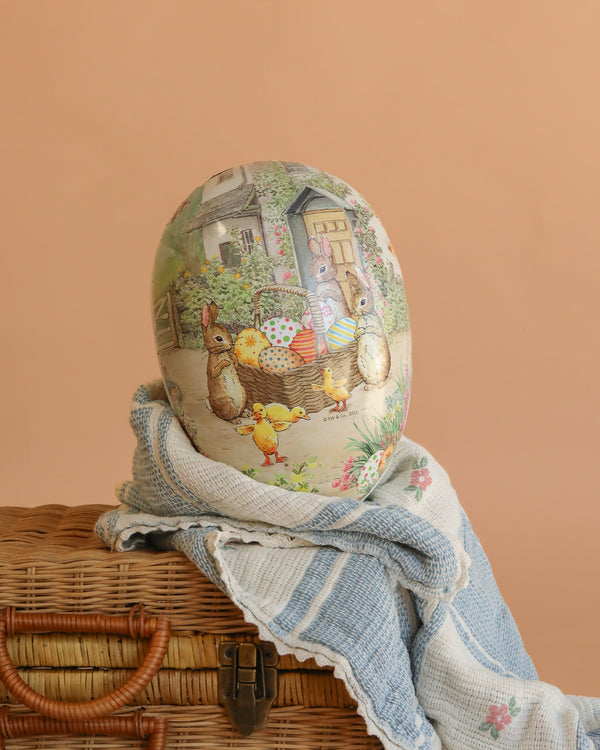 An extra large Cardboard Beatrix Potter Egg (Bunnies With Basket Of Eggs) sits atop a blue and white cloth in a wicker basket, displayed against a peach-toned background.