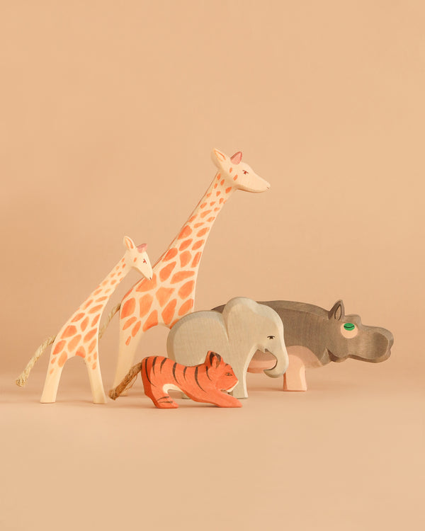 Five painted wooden animal figures—a giraffe, a calf, a tiger, an elephant, and a hippopotamus—stand side by side on a neutral background. The Ostheimer Handmade Wooden Safari Animals Set features these handcrafted toys made from sustainably sourced materials, blending timeless design with eco-friendly craftsmanship.