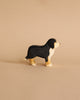 A small, wooden Ostheimer Bernese Mountain Dog with a black body, white chest, and tan feet stands on a neutral beige background. Made in the style of Ostheimer wooden toys, this piece appears to be hand-carved and painted using sustainable materials, capturing its form in a simple, artistic style.