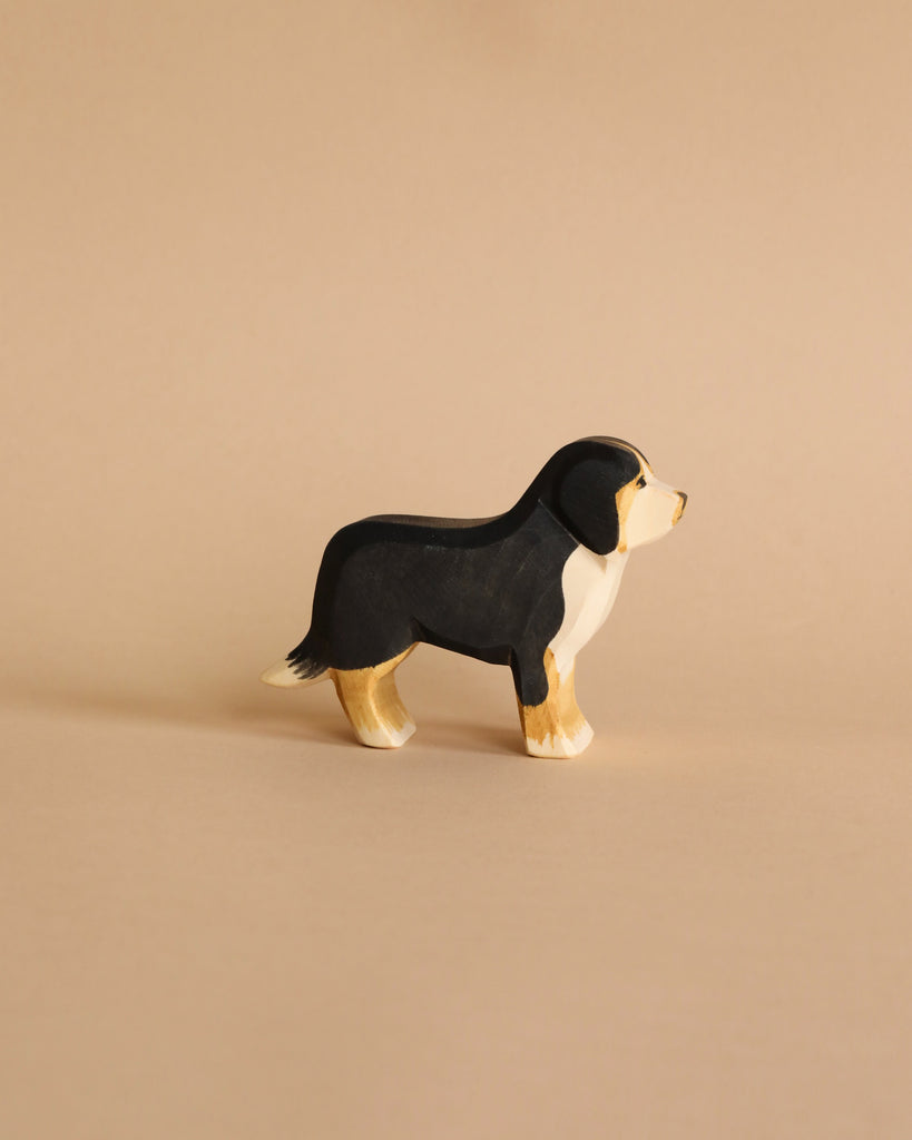 A small, wooden Ostheimer Bernese Mountain Dog with a black body, white chest, and tan feet stands on a neutral beige background. Made in the style of Ostheimer wooden toys, this piece appears to be hand-carved and painted using sustainable materials, capturing its form in a simple, artistic style.