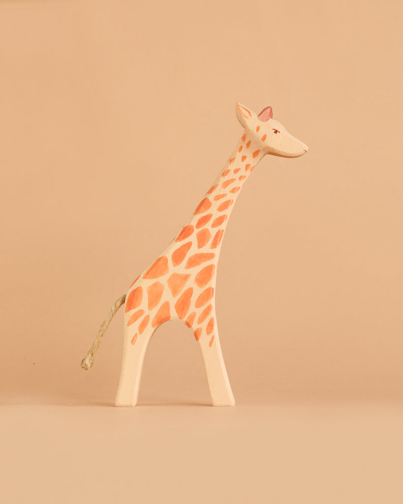 A handcrafted wooden giraffe figurine from the Ostheimer Handmade Wooden Safari Animals Set, featuring orange spots, stands on a plain beige surface against a matching beige background, reflecting the timeless quality and sustainable materials characteristic of Ostheimer toys.