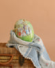 A large Cardboard Beatrix Potter Egg featuring the Rabbit Family in the Garden rests on a blue and white textured cloth atop a woven wicker basket, set against a plain peach background.