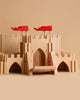 The Holztiger - Knight's castle is a handcrafted wooden European playset featuring red flags, a drawbridge, arched doorways, and battlements. Set against a beige background, its smooth construction makes it ideal for imaginative play.