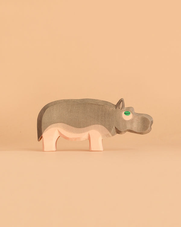 The Ostheimer Hippopotamus is a simple wooden toy featuring gray and pink colors, standing on a beige background. Handcrafted in the classic Ostheimer style, it has a green eye and minimal features, giving it a playful, minimalist appearance that encourages imaginative play.