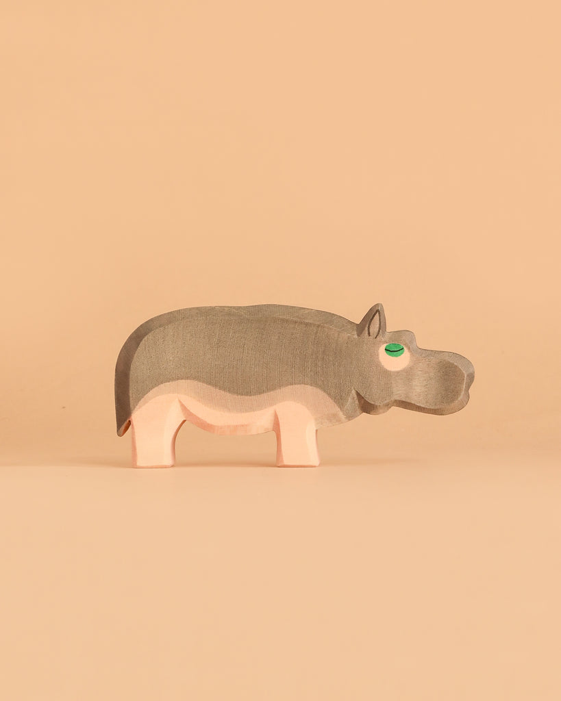 The Ostheimer Hippopotamus is a simple wooden toy featuring gray and pink colors, standing on a beige background. Handcrafted in the classic Ostheimer style, it has a green eye and minimal features, giving it a playful, minimalist appearance that encourages imaginative play.