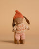 The Maileg Winter Bunny, Micro - Brown - Rose wears a knitted orange hat, cream and orange sweater, and pink checkered shorts—ideal for miniature dollhouse play or pairing with Maileg accessories against a beige background.
