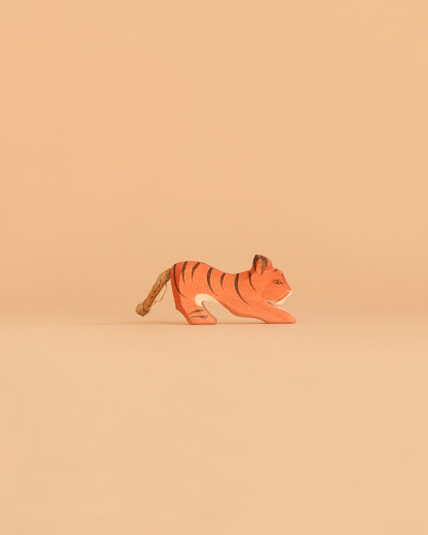 A small Ostheimer Tiger Lurking figurine, crafted from wood and painted in orange with black stripes, stands on a plain beige surface against a solid beige background—a charming example of the timeless quality and craftsmanship of Ostheimer wooden toys.