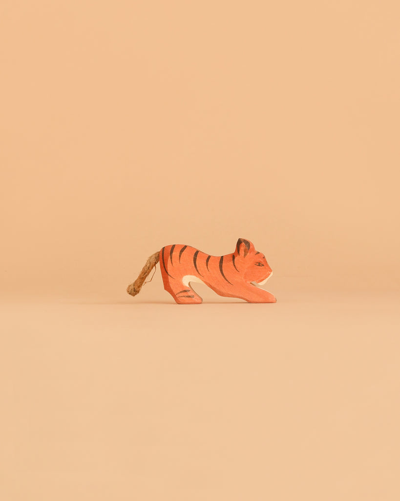 A small Ostheimer Tiger Lurking figurine, crafted from wood and painted in orange with black stripes, stands on a plain beige surface against a solid beige background—a charming example of the timeless quality and craftsmanship of Ostheimer wooden toys.