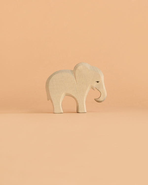 The Ostheimer Small Elephant – Eating stands on a solid peach background, facing right, featuring simple carved details and a minimalist design—perfect for imaginative play and evoking the charm of handcrafted wooden toys.