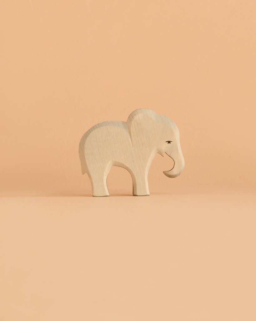 An elegant elephant figurine from the Ostheimer Handmade Wooden Safari Animals Set, inspired by traditional handcrafted wooden toys, stands on a simple peach-colored background.