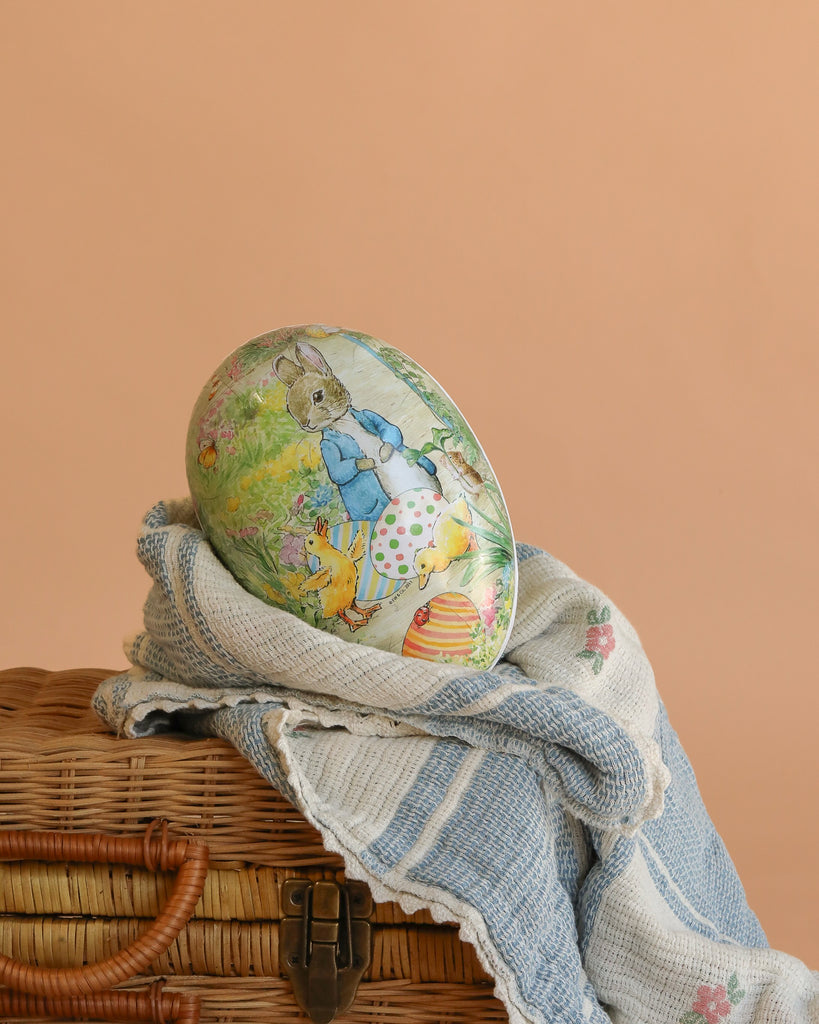 A large handcrafted Cardboard Beatrix Potter Egg (Rabbit with Ducklings) rests on a blue-and-white striped cloth atop a wicker basket, set against a peach-colored background.