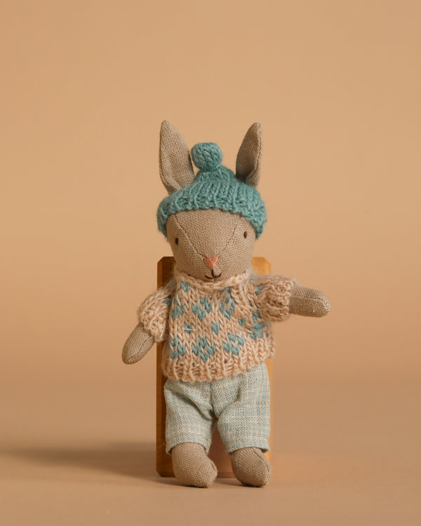 The Maileg Winter Rabbit, Micro - Sand - Mint wears a blue knit hat, beige and blue sweater, and light blue pants, sitting on a wooden chair against a beige background—ideal for dollhouse displays or Maileg accessories.