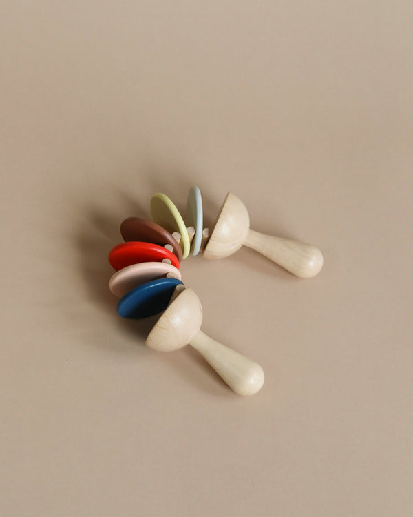 Two sustainably made Clatter Percussion Toys with colorful, pastel-colored bands around the tops positioned in a cross shape on a beige background.