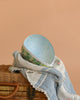 A Cardboard Beatrix Potter Egg, Large (Morning at Ginger & Pickles’ Shop), reminiscent of pastel German Easter eggs, sits on a woven picnic basket partly covered by a blue and white striped blanket with pink flower accents.