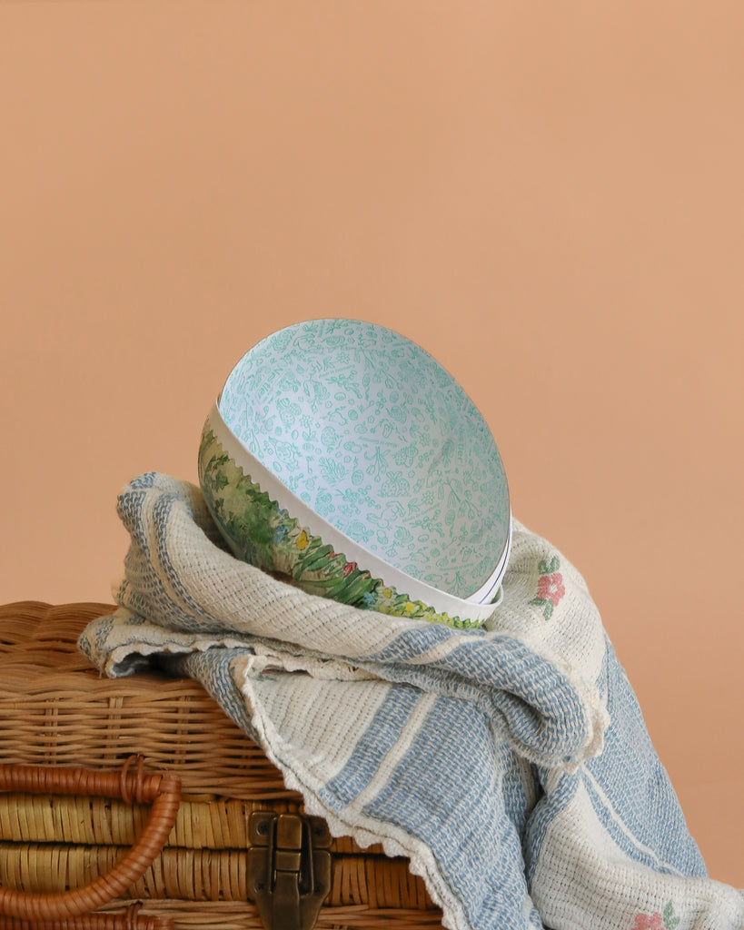 A Cardboard Beatrix Potter Egg, Large (Morning at Ginger & Pickles’ Shop), reminiscent of pastel German Easter eggs, sits on a woven picnic basket partly covered by a blue and white striped blanket with pink flower accents.