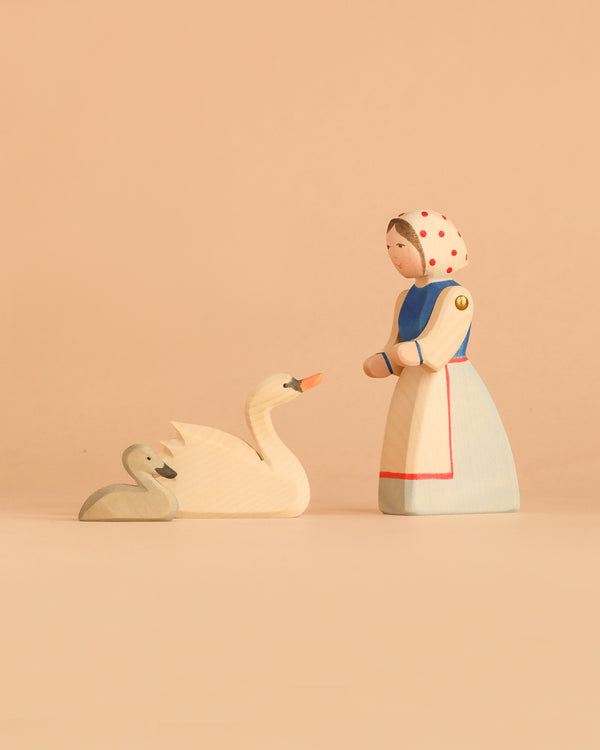 The Ostheimer Handmade Wooden Swan Girl Set features a beautifully handcrafted wooden figurine of a woman in a white dress and headscarf, standing before two wooden swans—one large and one small—set against a plain beige background. Inspired by classic Ostheimer toys and crafted from sustainably sourced materials, this set embodies timeless charm while reflecting care for the environment.