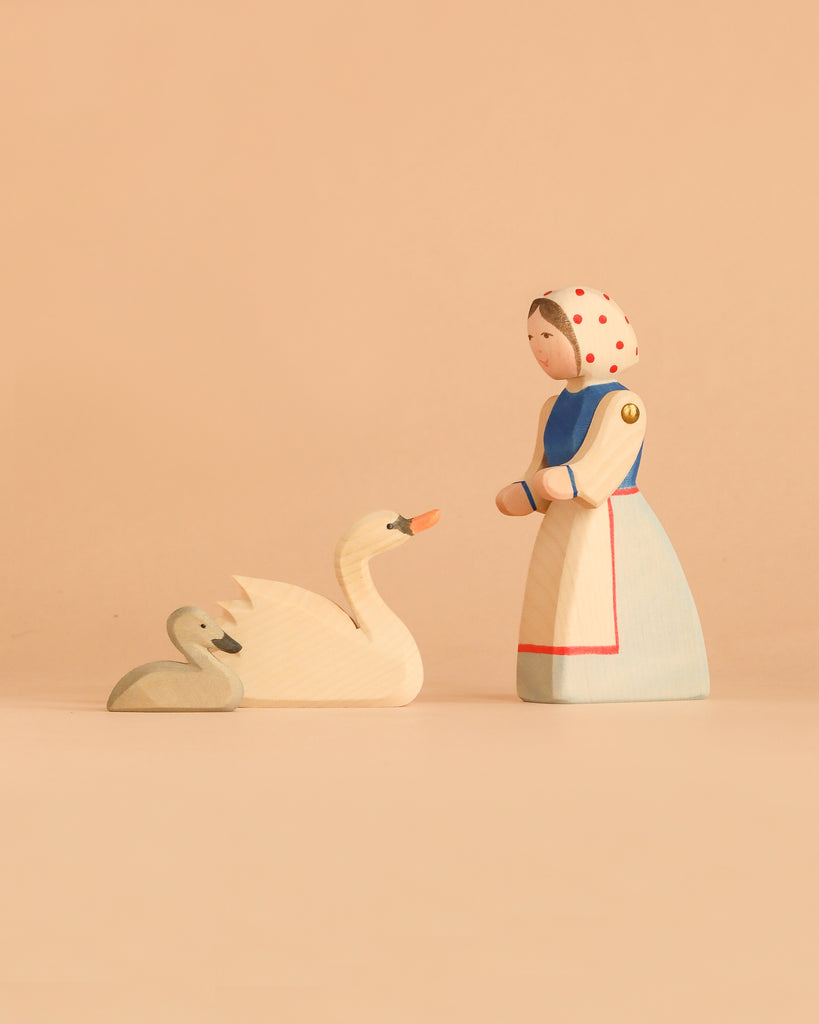 The Ostheimer Handmade Wooden Swan Girl Set features a beautifully handcrafted wooden figurine of a woman in a white dress and headscarf, standing before two wooden swans—one large and one small—set against a plain beige background. Inspired by classic Ostheimer toys and crafted from sustainably sourced materials, this set embodies timeless charm while reflecting care for the environment.