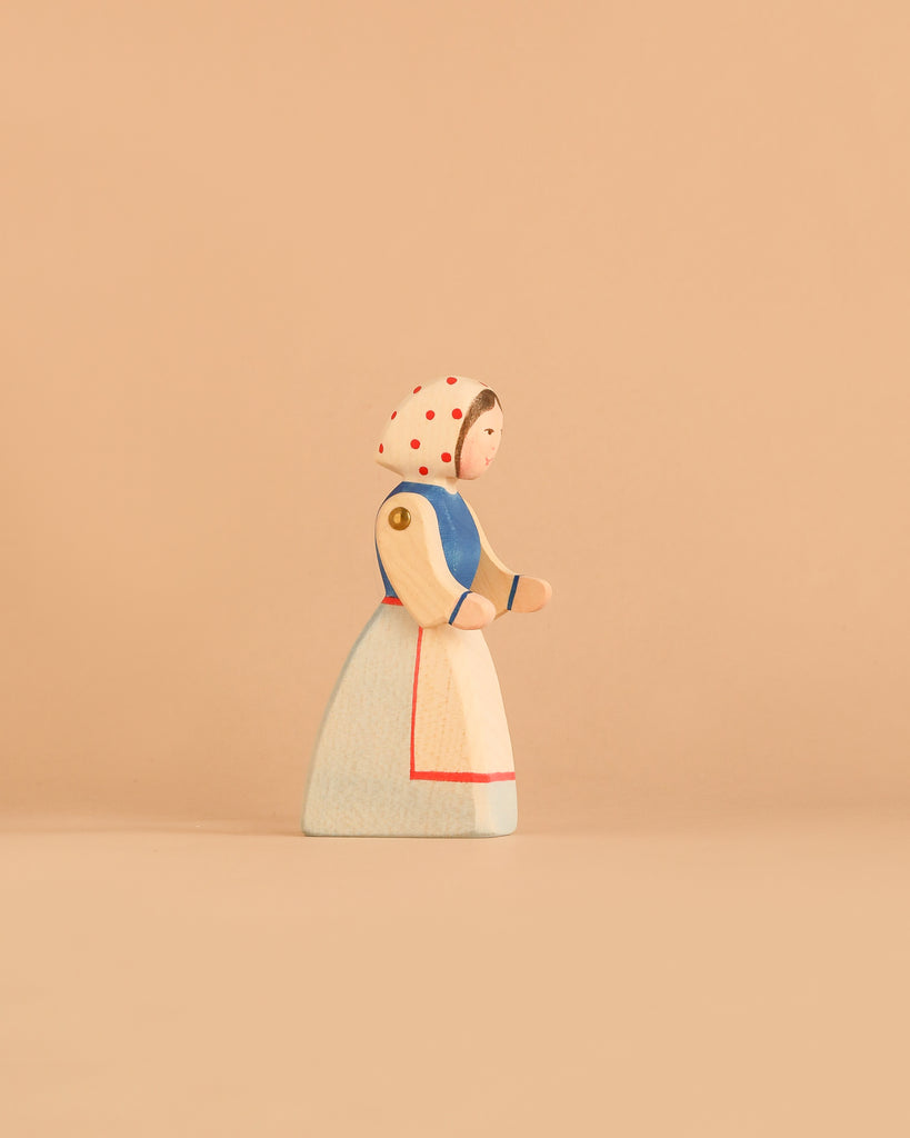 The Ostheimer Farm Wife is a handcrafted wooden figurine featuring a woman in a blue and white dress with a white bonnet covered in red dots, set against a plain beige background—an ideal collectible for those who appreciate sustainable wooden toys.