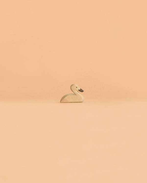 A small, light-colored handcrafted wooden Ostheimer Swan figurine is displayed on a smooth, peach-colored surface with a matching peach background.
