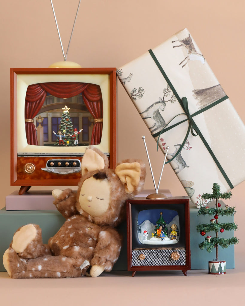 A cozy holiday scene featuring a plush deer toy, a Charlie Brown Christmas Music TV illuminating festive decorations, a small decorated tree, and a wrapped gift adorned with winter-themed illustrations softly playing a Christmas melody.