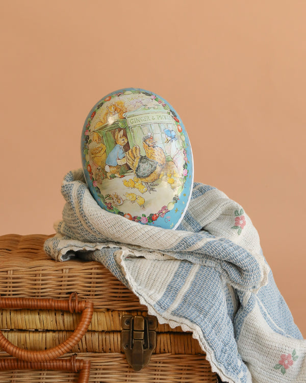 The Cardboard Beatrix Potter Egg, Large (Morning at Ginger & Pickles’ Shop), featuring a vintage illustration, sits on a blue and white striped cloth atop a wicker basket against a light peach background.